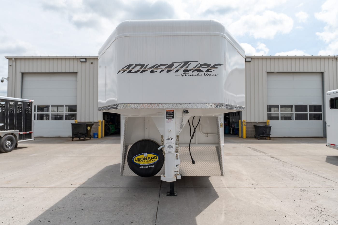 Front view of a white gooseneck trailer with 'ADVENTURE' branding, parked in a service yard in front of a building with two bay doors.