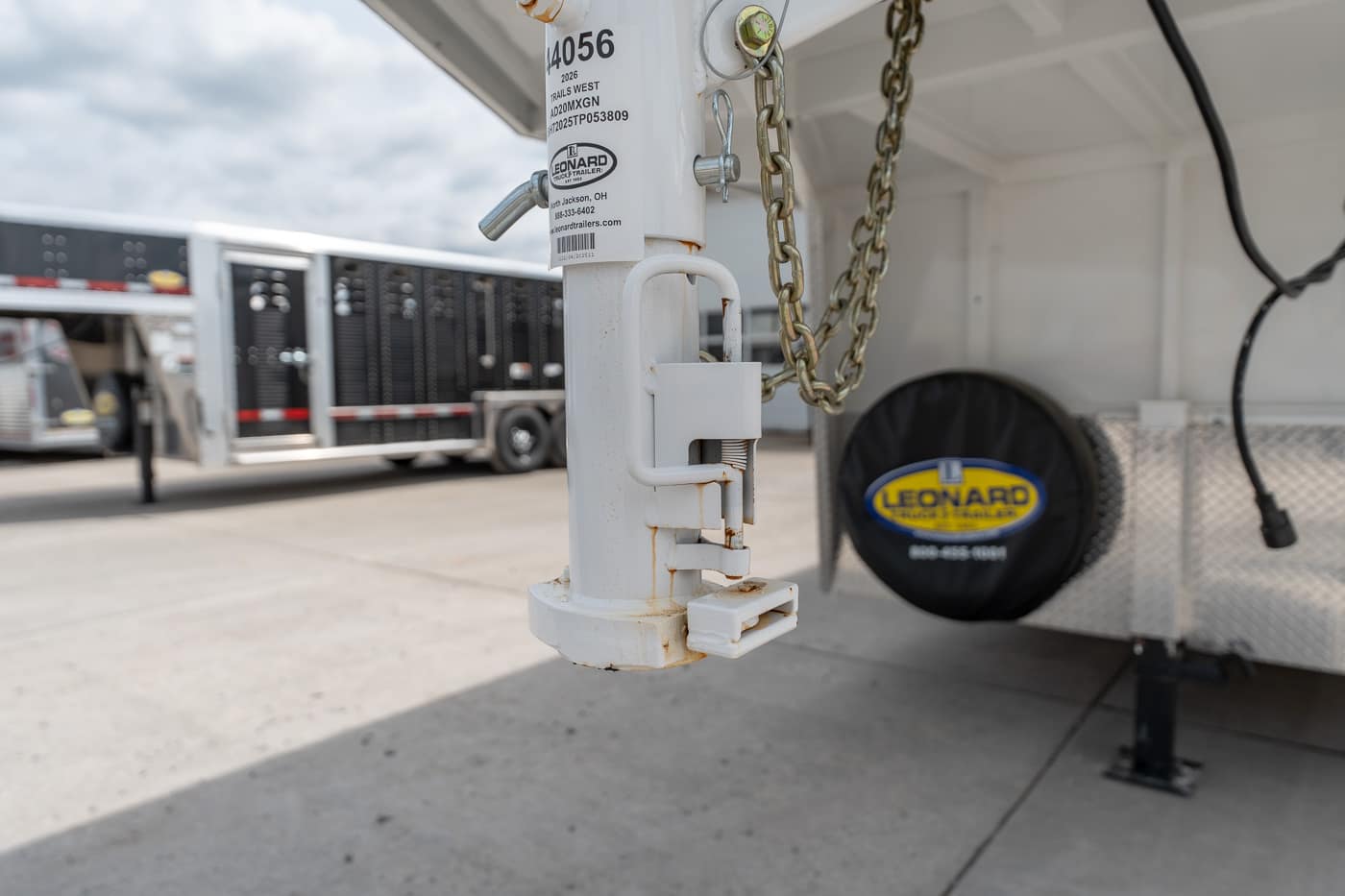Close-up of a white trailer hitch with a locking latch and chain, with a Leonard trailer and spare tire in the background.