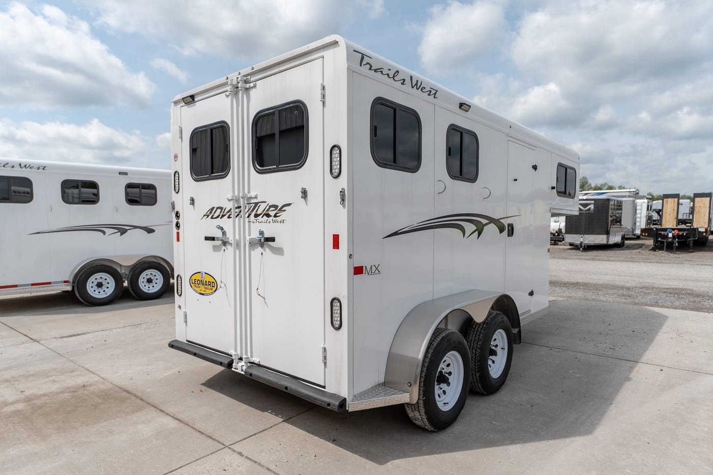 White dual-axle horse trailer with split rear doors and small windows, parked in a lot with other trailers under a partly cloudy sky. It has decorative striping and a Trails West branding on the top edge.