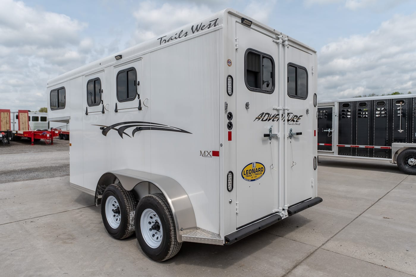 White Trails West two‑axle horse trailer with rear double doors and side windows, parked on a concrete lot; features decorative black graphics and a Leonard trailer sticker on the back door.