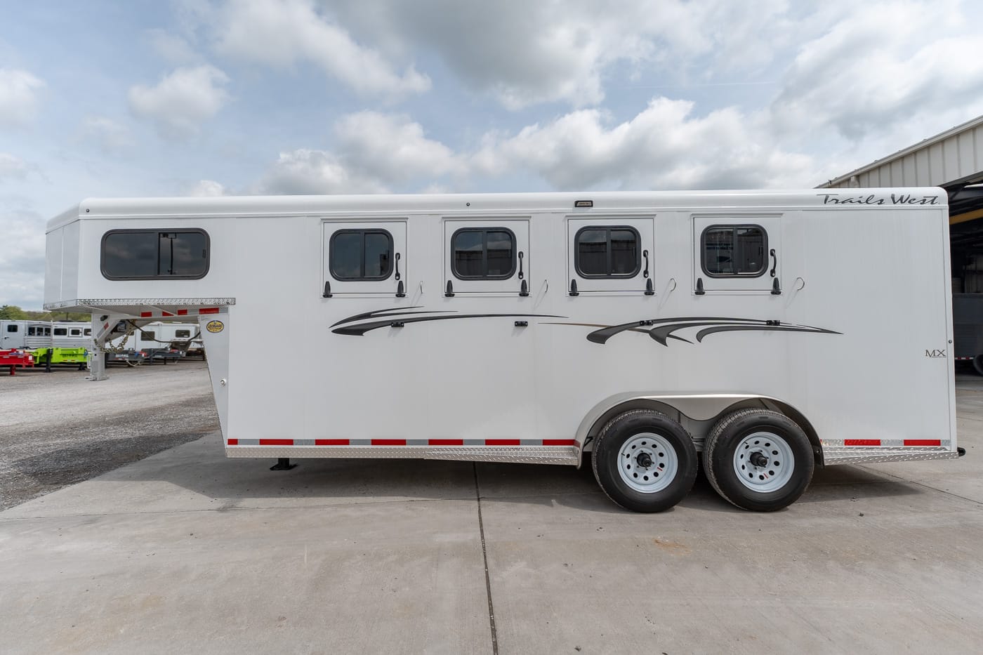 White horse trailer with four side windows and decorative black graphics, parked on a concrete lot under a cloudy sky.