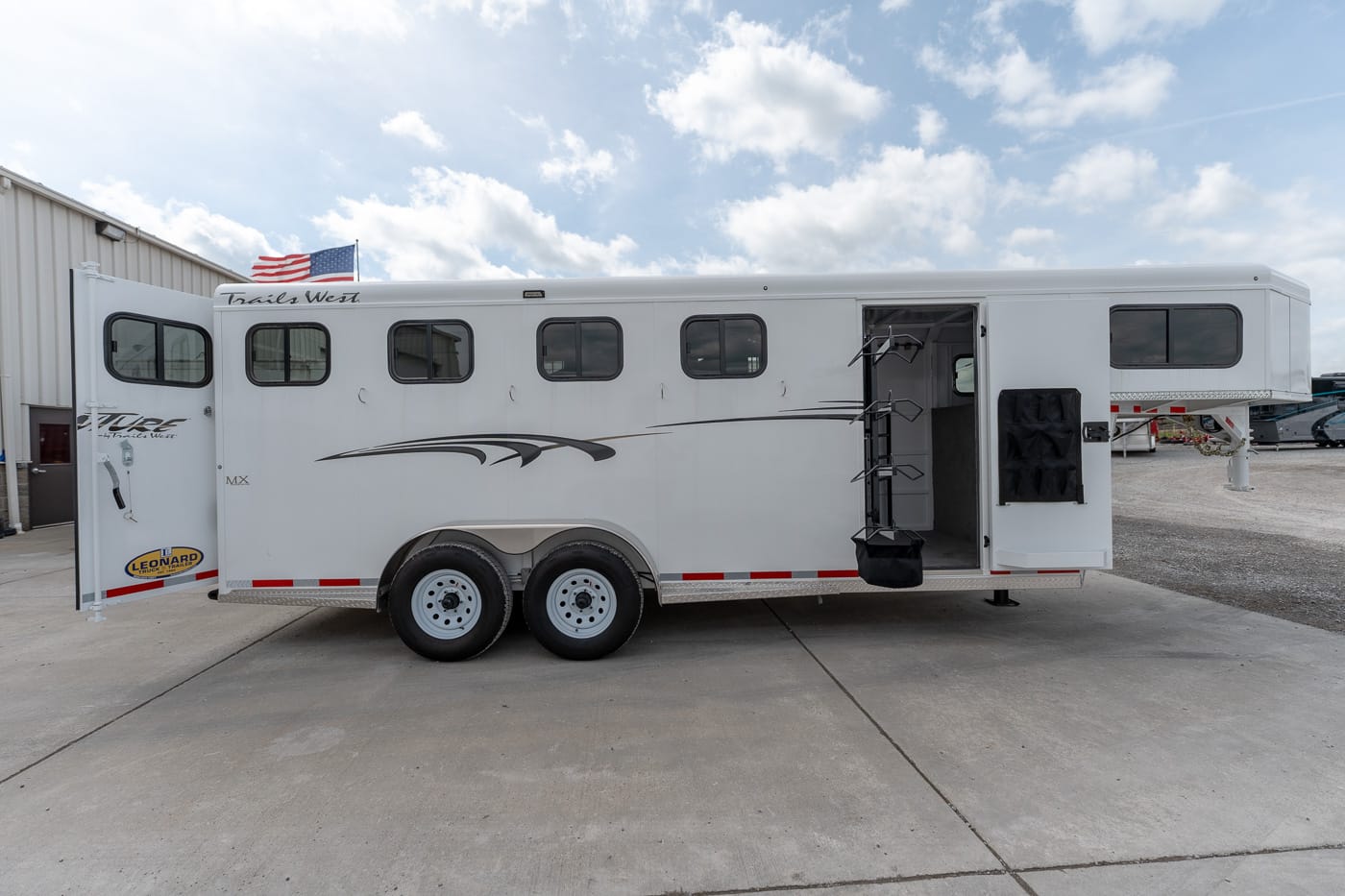 White Trails West horse trailer with multiple side windows, open side entry, and exterior tack racks parked on a concrete lot under a blue sky with clouds