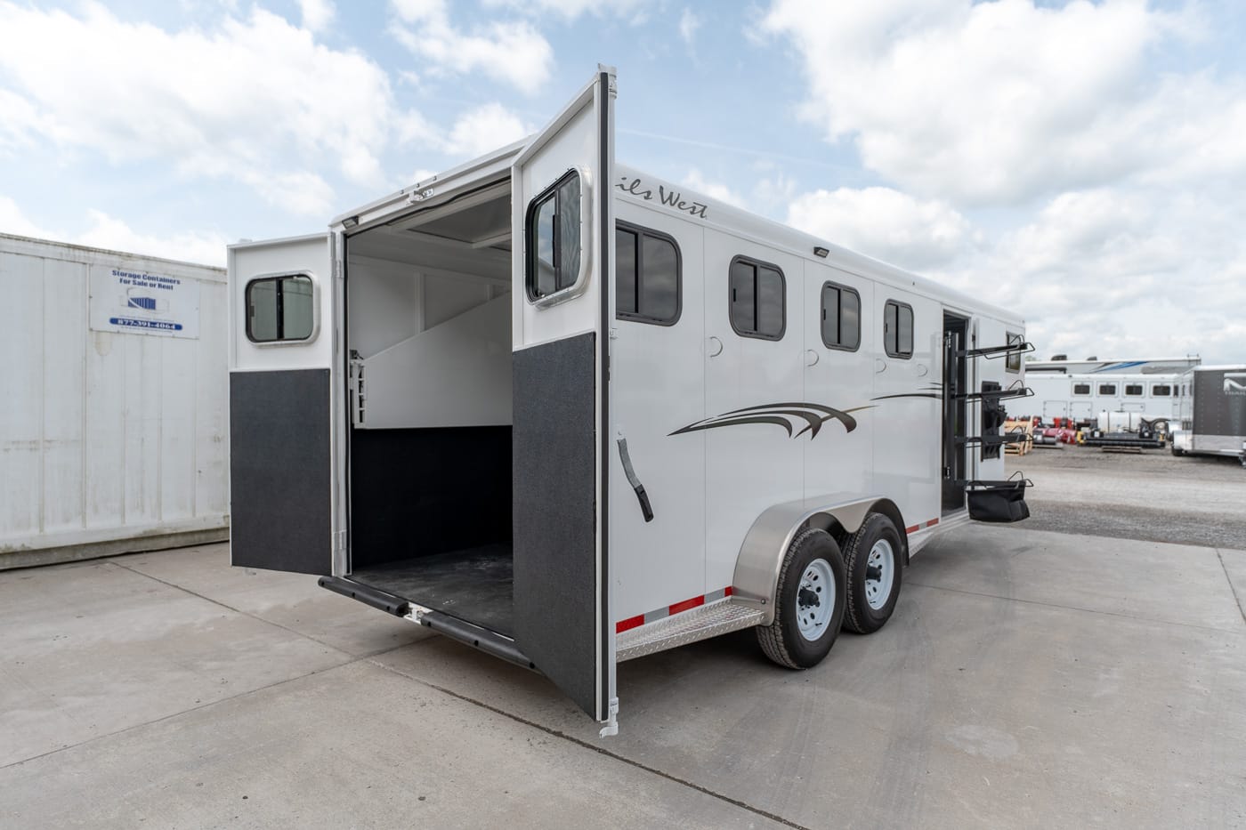White horse trailer with open rear ramp showing interior stalls and divider, parked on a concrete lot under a cloudy sky