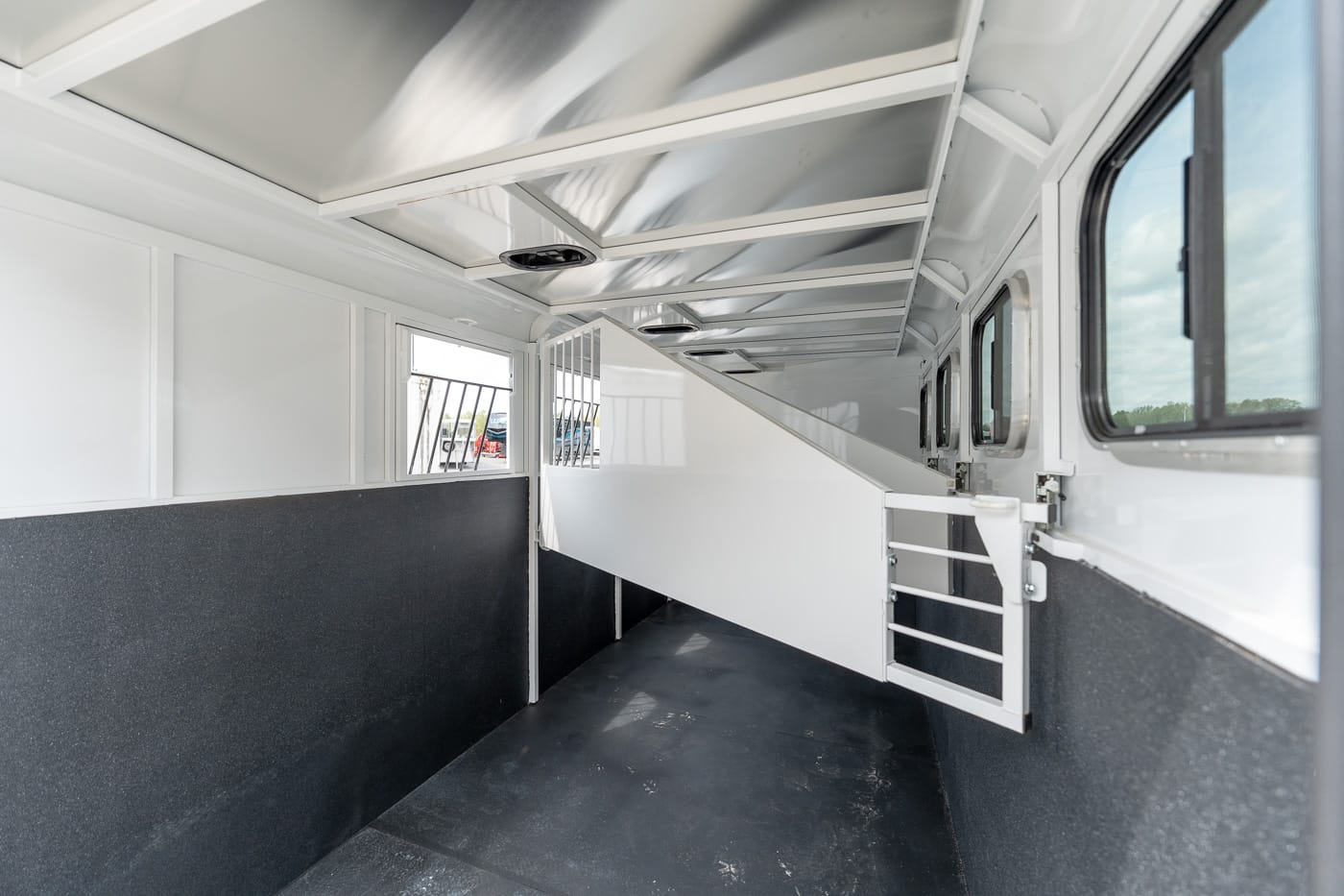 Interior of a white horse trailer with a central divider forming a stall, dark non-slip floor, and side windows with bars on the left/right.
