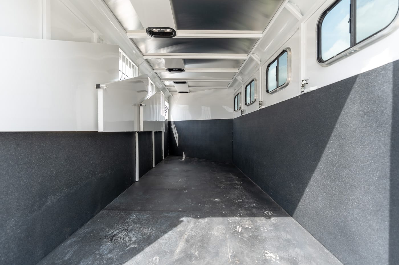 Empty horse trailer interior with white upper walls and dark lower panels, stall dividers on the left and windows along the right wall.