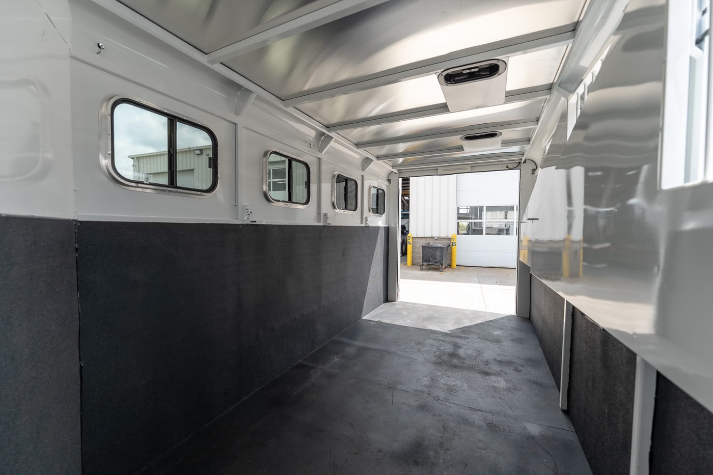 Interior of an empty white transport trailer with dark padded walls and small side windows, open at the rear to loading area outside.