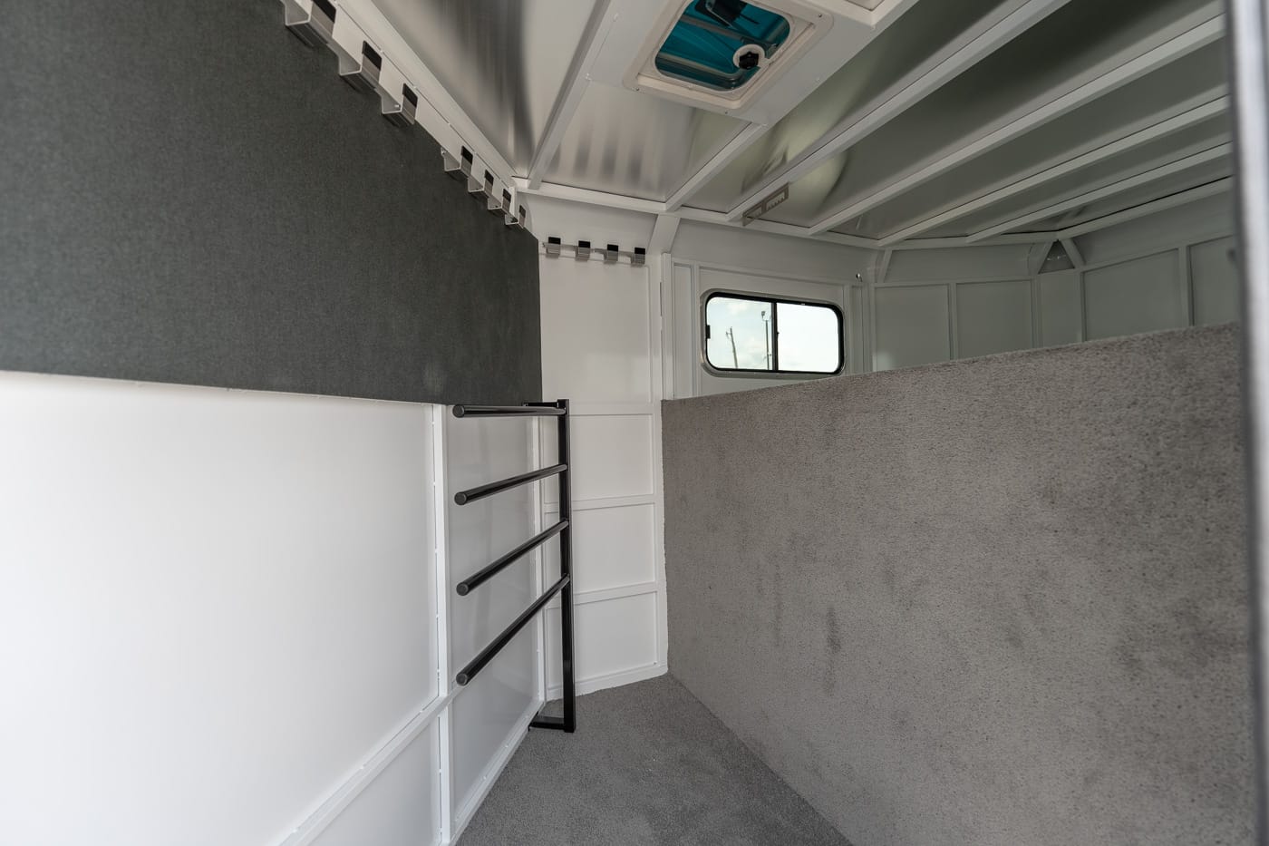 Interior of a white horse trailer with gray carpeting, black divider rails along the left wall, and a small window at the far end.