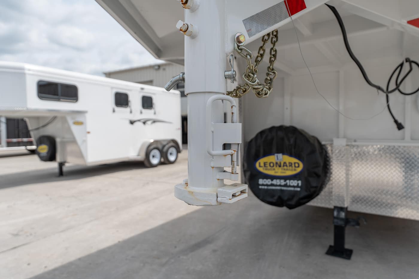 Close-up of a white trailer hitch with a padlock and chain, mounted on a gooseneck trailer in a parking lot.