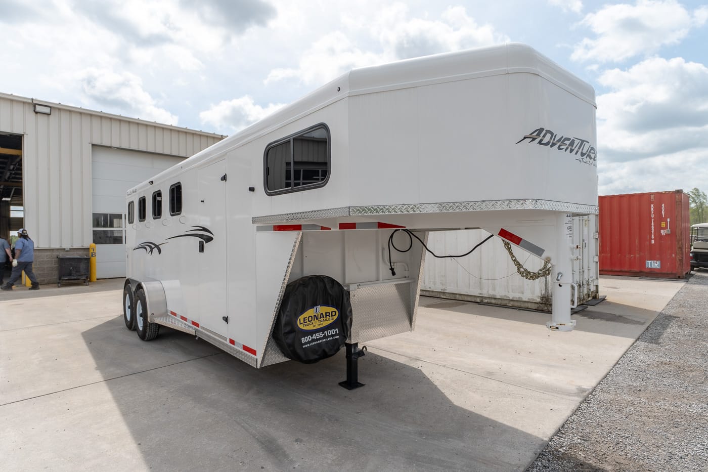 White gooseneck horse trailer parked outside a workshop, with spare tire cover and refuge jack at the front
