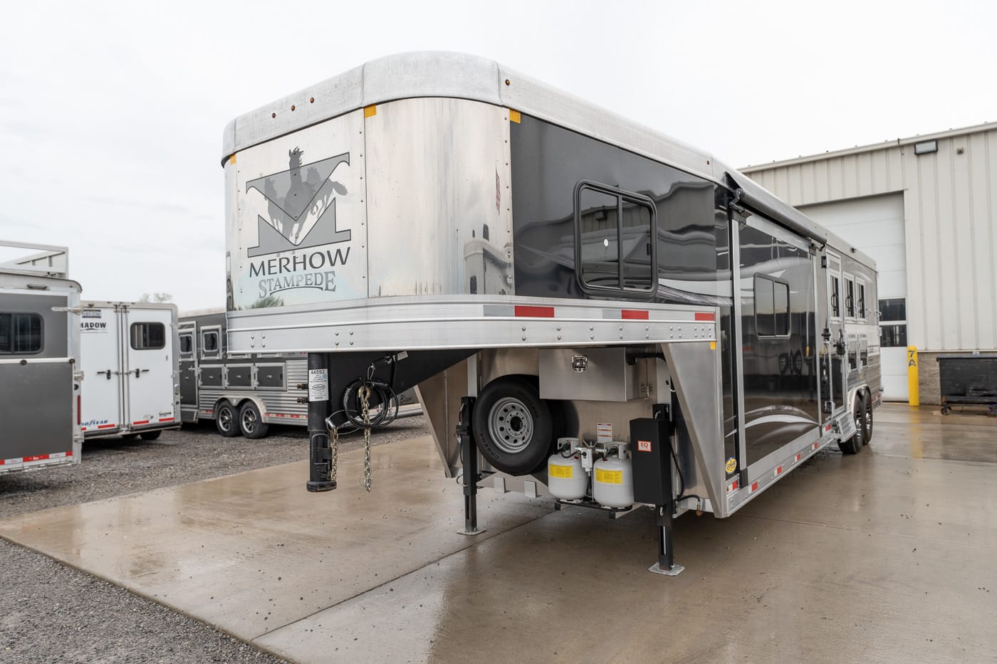 Silver Merhow Stampede horse trailer parked on wet concrete, showing exposed propane tanks and spare tire undercarriage.