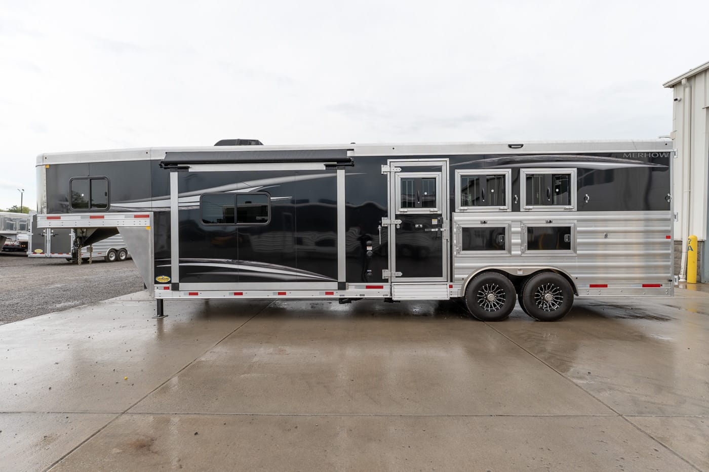 Two-tone black and silver horse trailer with multiple windows and a side door, parked on a wet concrete lot.