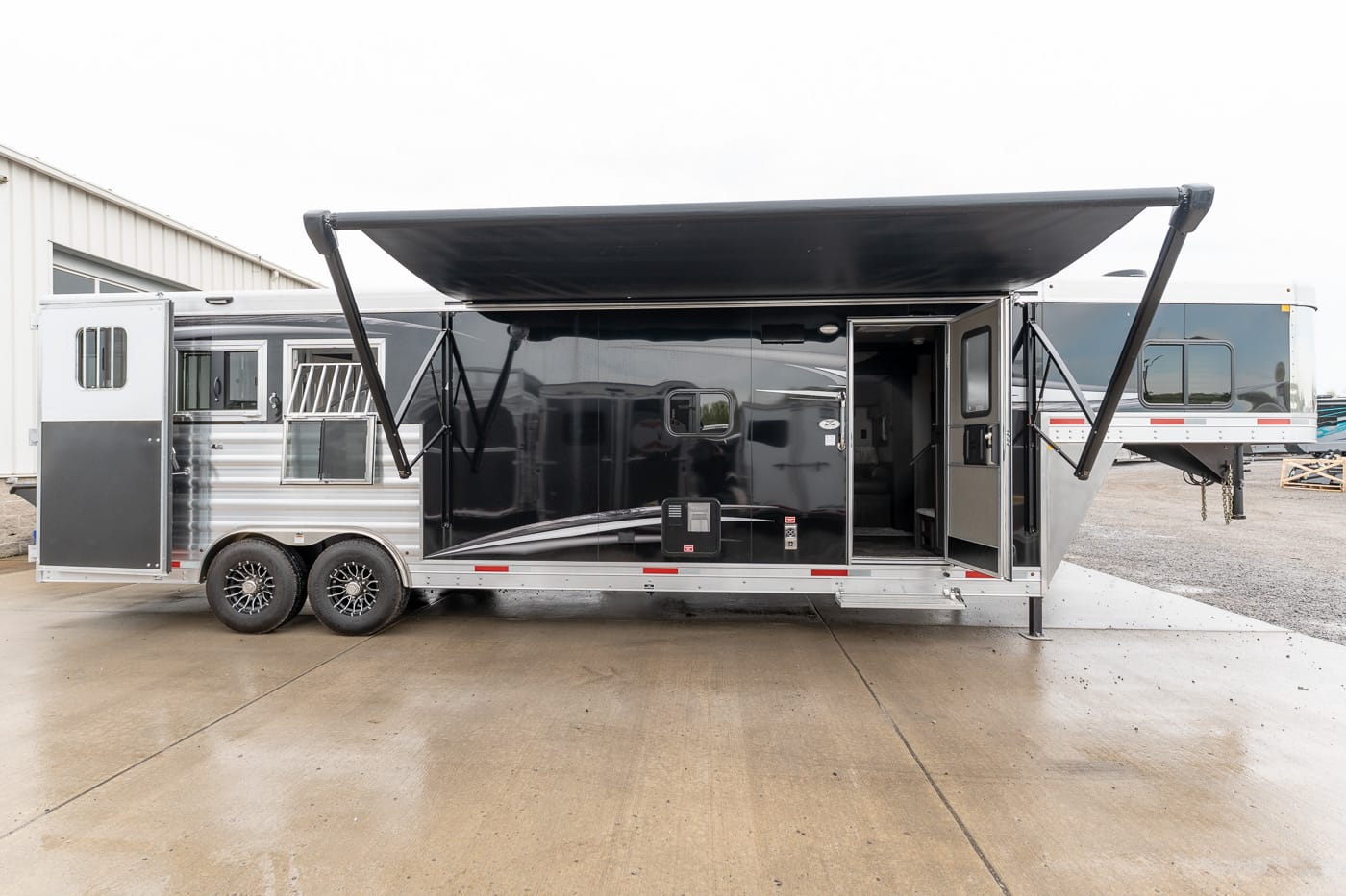 Black and silver gooseneck travel trailer with extended awning and open entry door on a concrete pad.
