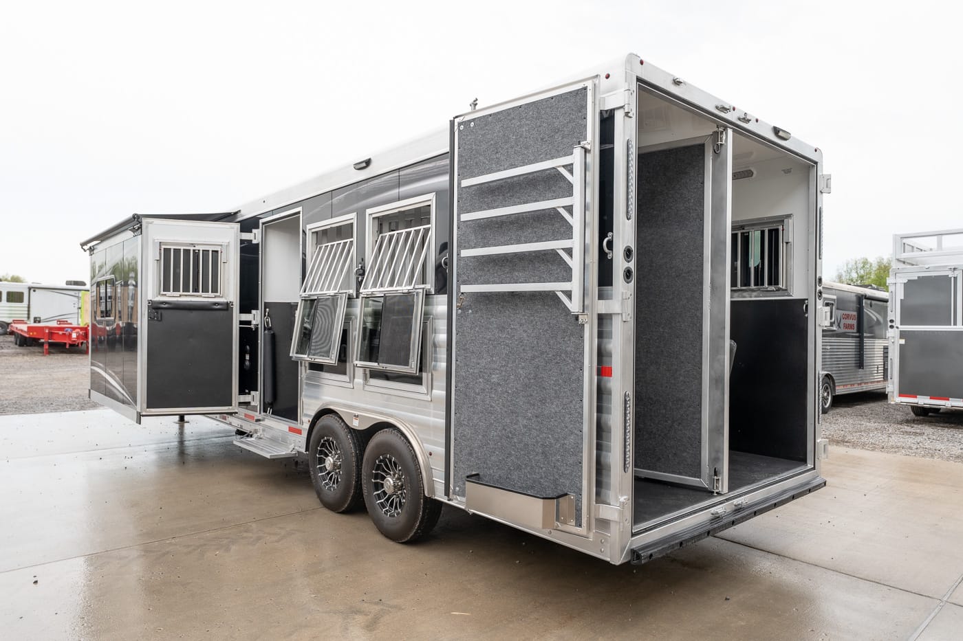 Aluminum horse trailer with open rear ramp, tack storage doors, and barred windows on its side. Two-axle design visible on a concrete lot.