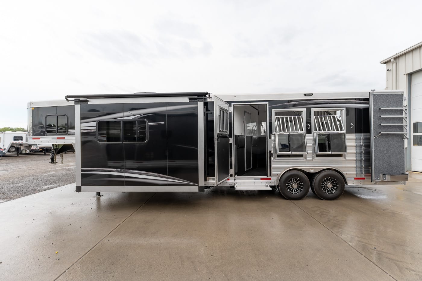 Two-axle black and silver horse trailer with the side door open, revealing interior stalls and barred windows.