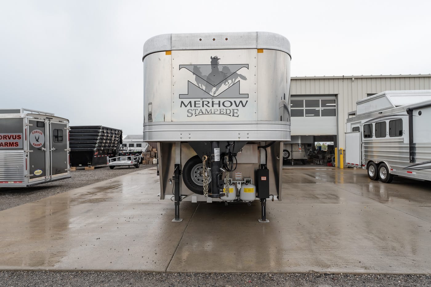 Front view of a large silver Merhow Stampede horse trailer parked in a lot, with other trailers and a workshop in the background.