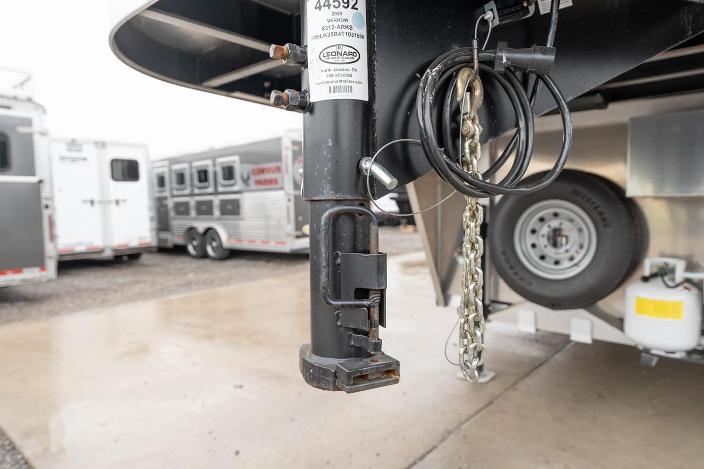 Close-up of a trailer jack and safety chain hanging from a gooseneck trailer hitch, with electrical cables nearby.