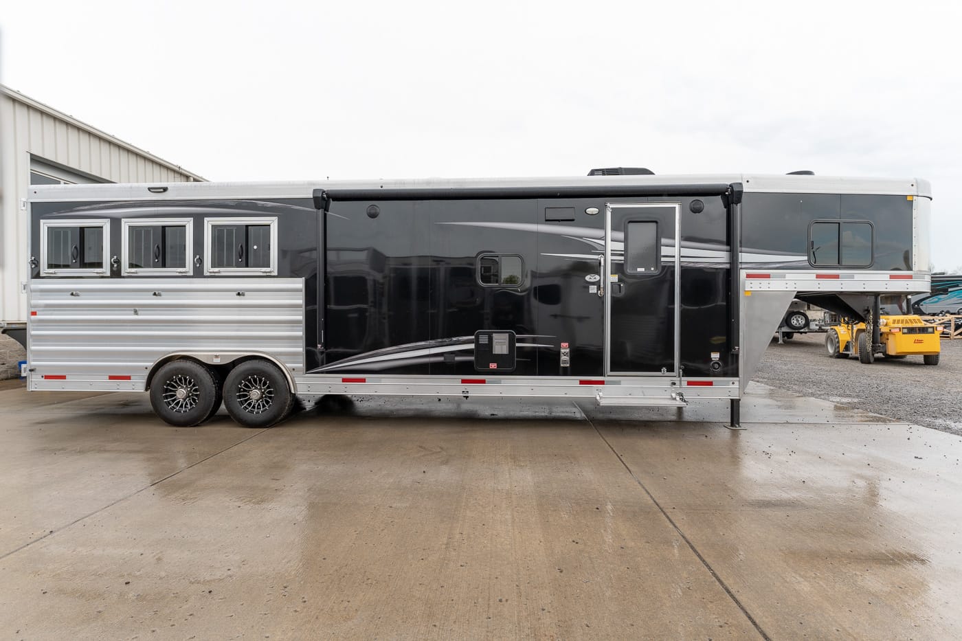 Long black and silver horse trailer with three side windows, a raised gooseneck, and tandem axles parked on wet concrete.