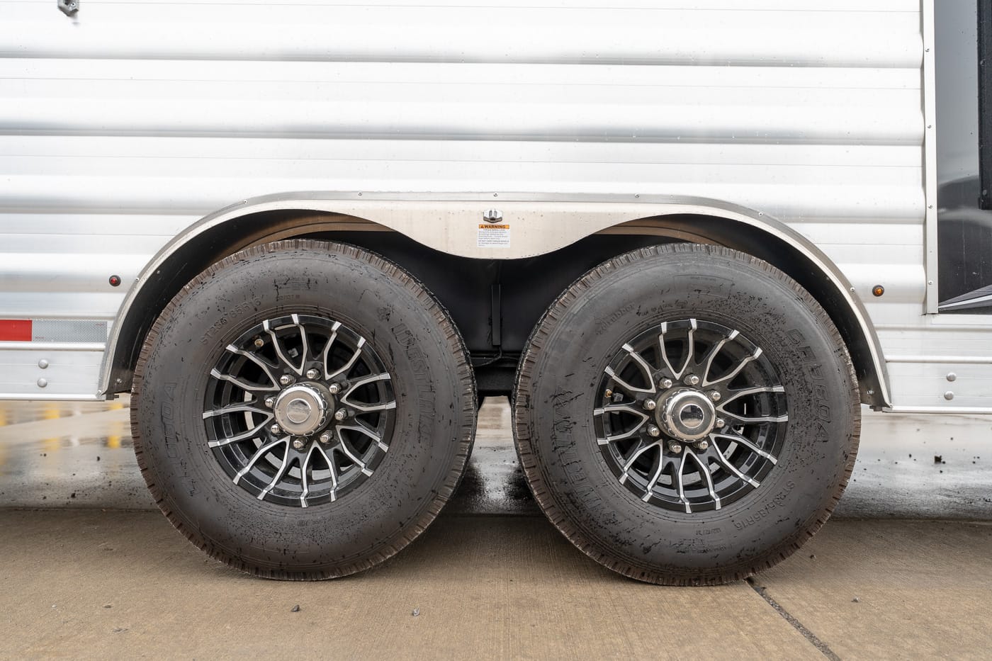 Dual-axle trailer wheels with black spoked rims on a silver trailer. Two large black tires and alloy wheels of a tandem trailer under a metal fender on a silver camper body.