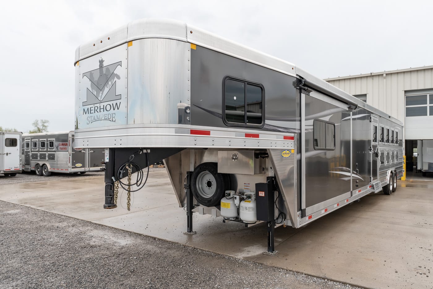Silver Merhow Stampede horse trailer with gooseneck front, windows along the side, spare tire and propane tanks, parked outside a service building.