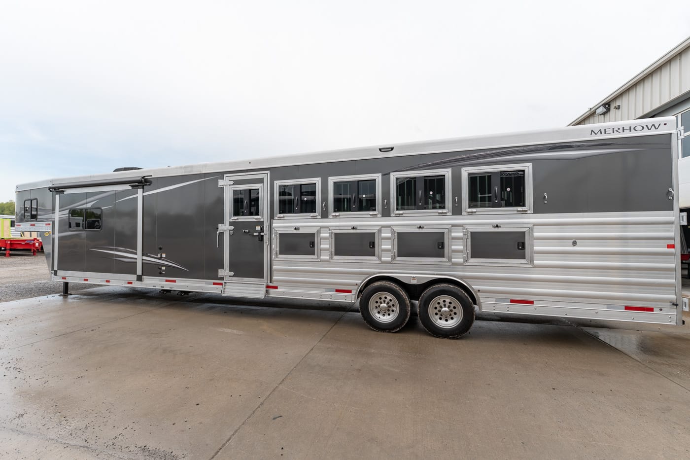 Grey and silver horse trailer with multiple stalls/windows and tandem axles, parked on concrete lot (Merhow branding visible).