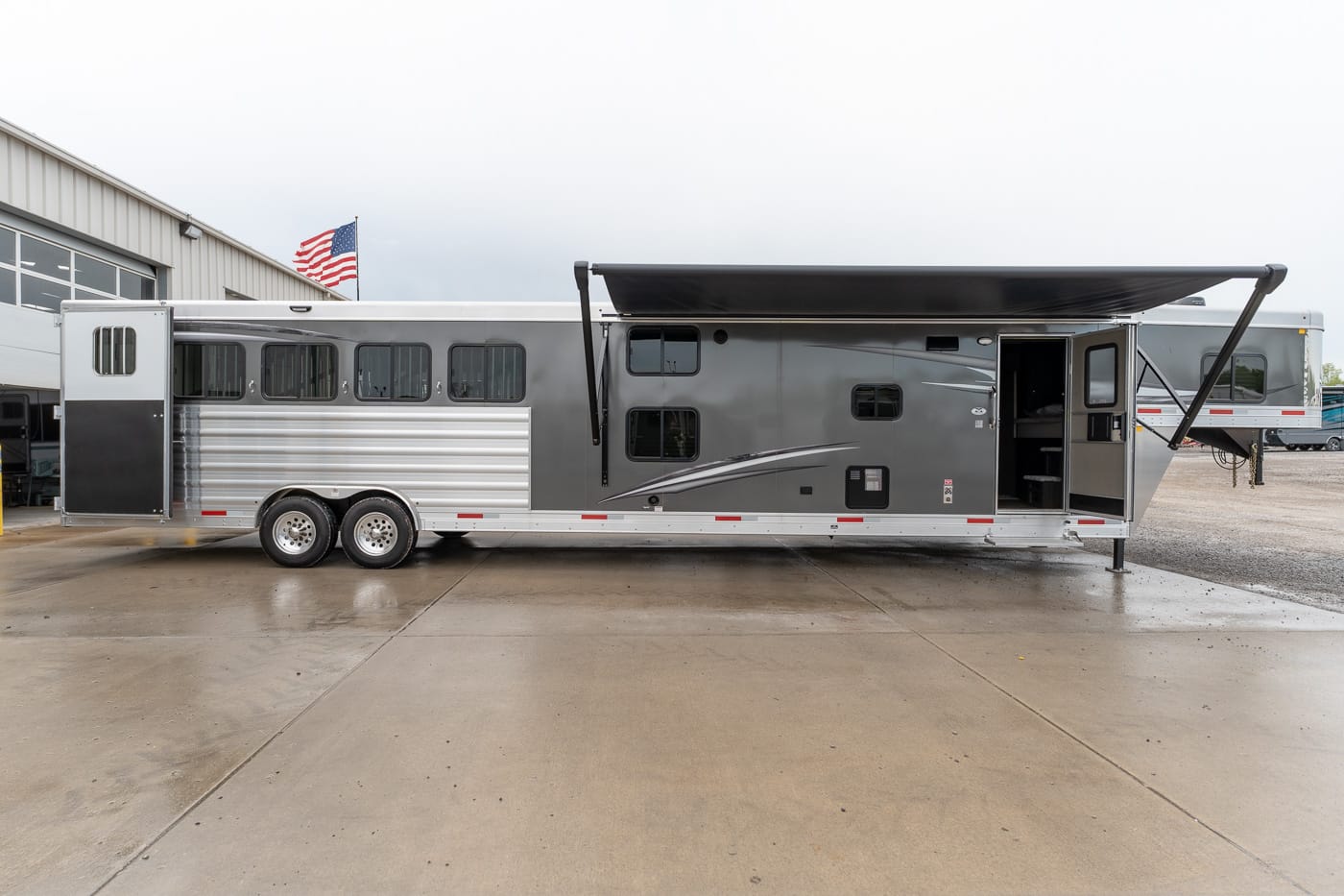 Grey and silver multi-section horse trailer with windows along the side, a large black retractable awning extended above the entry, and an open doorway showing interior compartments; parked on a wet concrete lot with an American flag in the background.