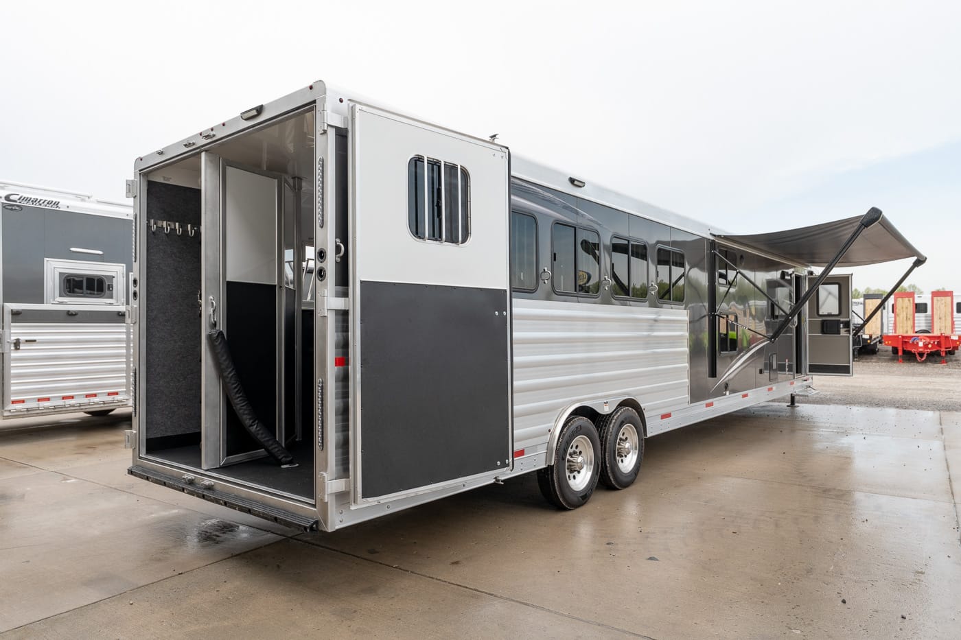 Silver tandem-axle trailer with open rear ramp, showing interior partition and horse stalls; exterior awning extended on a wet lot.