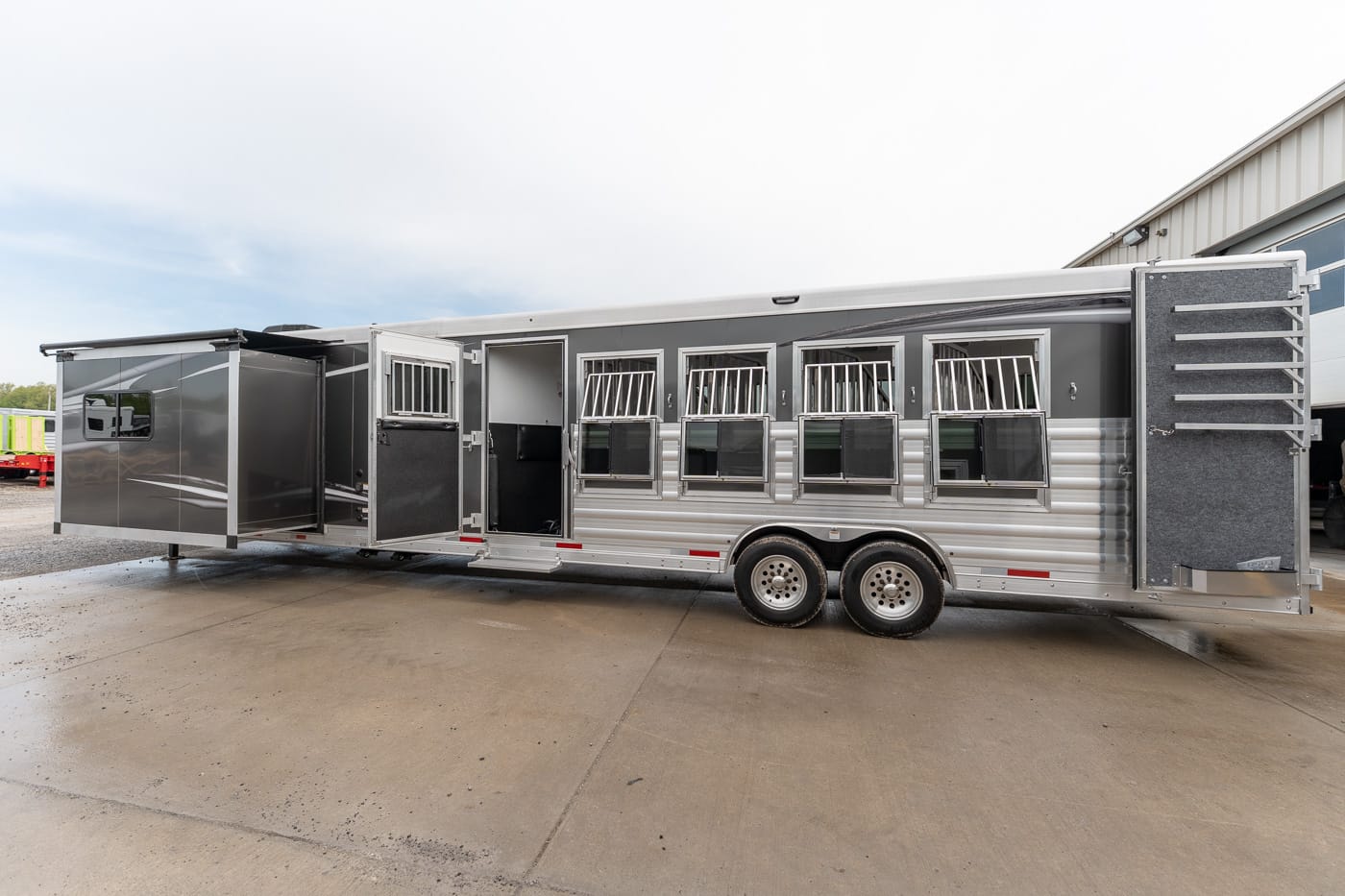 Silver aluminum multi-stall horse trailer with several barred windows and a side entry door, resting on a wet concrete lot with the rear ramp extended.