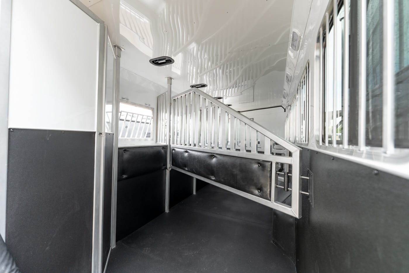 Interior of an empty metal horse trailer with padded black walls and a slatted divider forming a ramp or stall barrier for horse loading/safety at the back of the trailer behind windows and aluminum framing.
