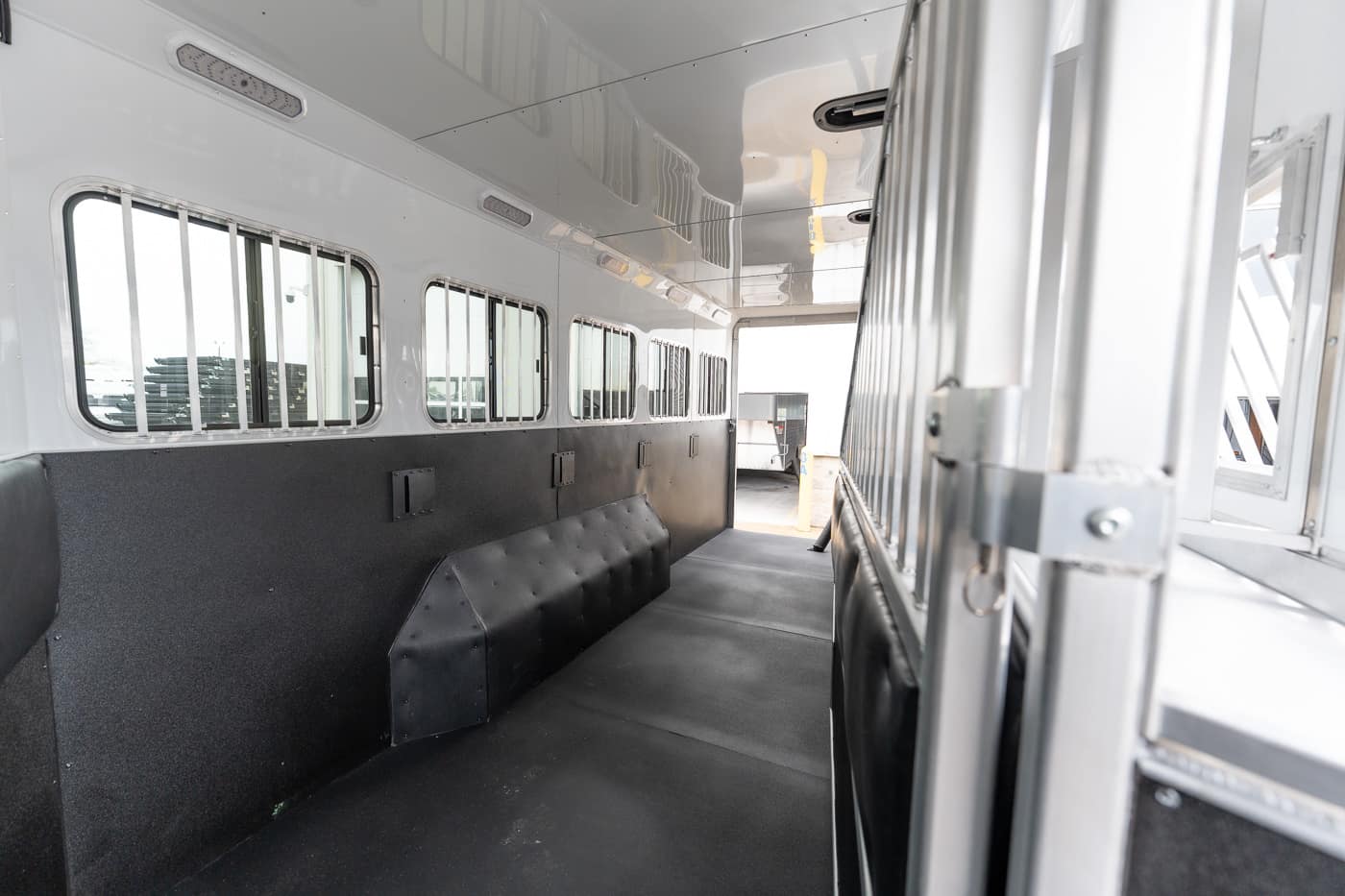 Interior of a white livestock trailer: lined with barred windows along the left wall, black padded partitions and a rubber floor, with a metal divider on the right and the rear door open to the outside light.