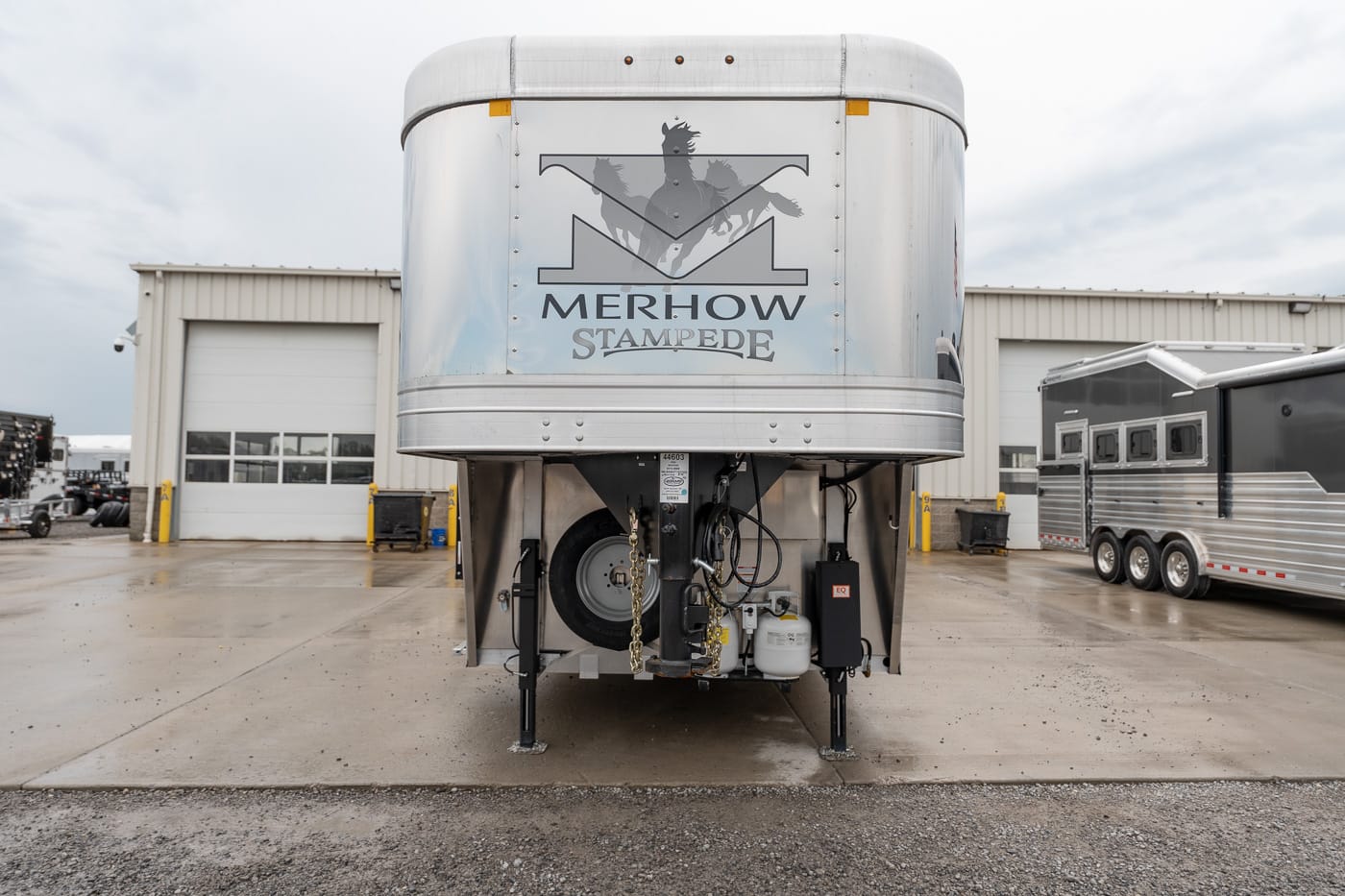 Silver horse trailer in a facility lot with a Merhow Stampede logo on the front panel; other trailers and a warehouse in the background.