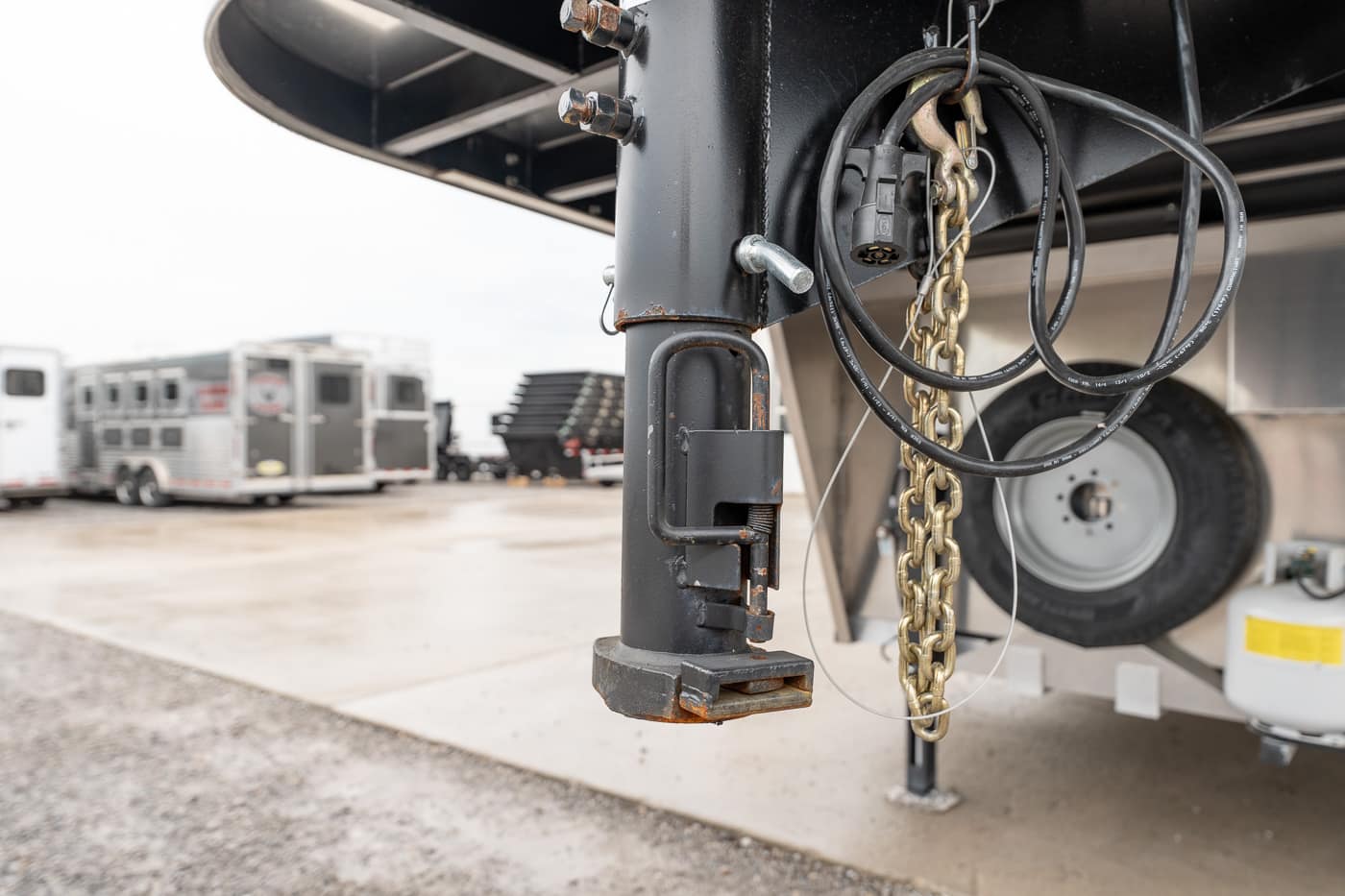 Close-up of a trailer jack and safety chains hanging from a black trailer, with a spare tire in the background on a wet lot.