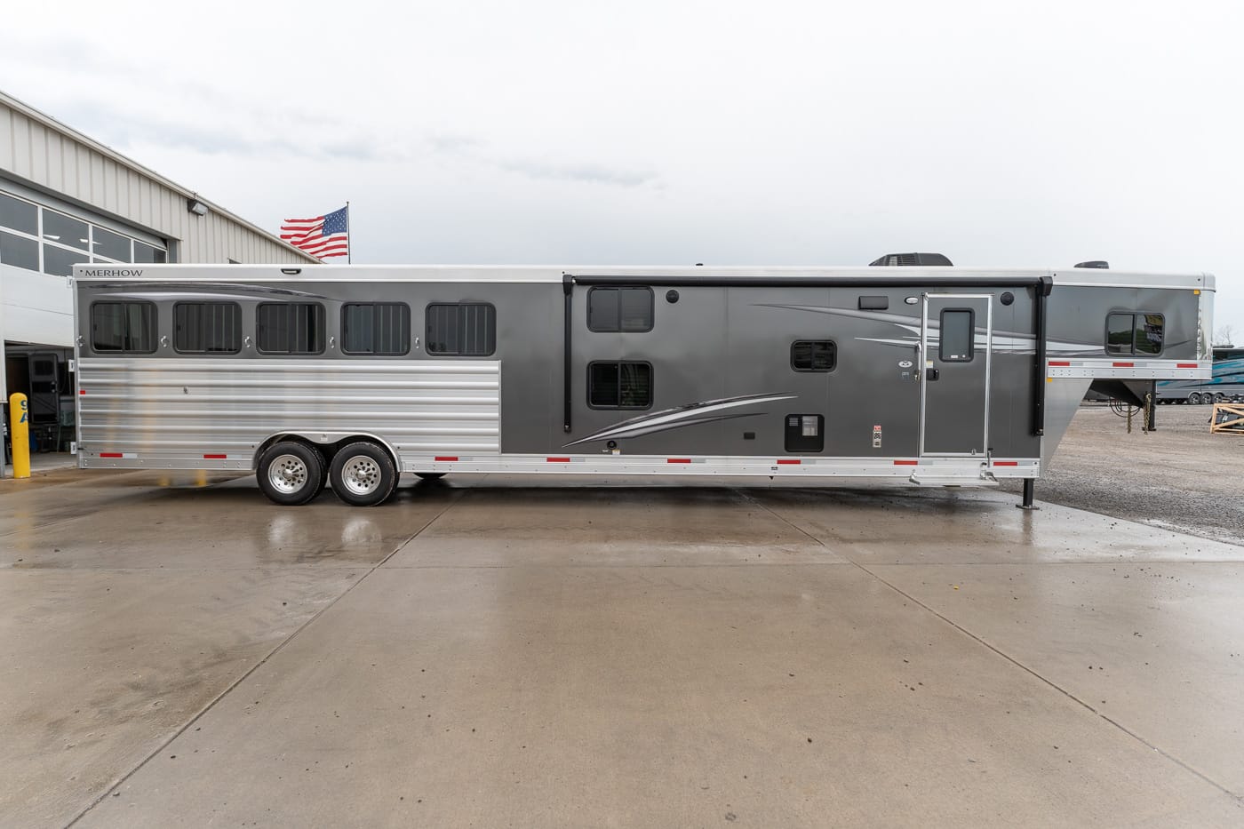 Long gray horse trailer parked on a concrete lot beside a building with an American flag in the background