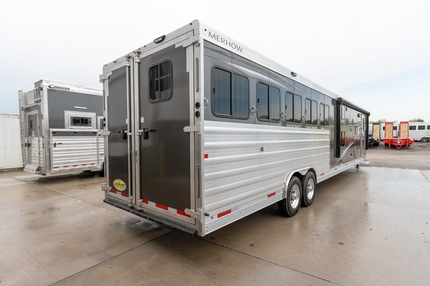Merhow horse trailer with silver aluminum siding, side windows, and rear double doors on a dealership lot.