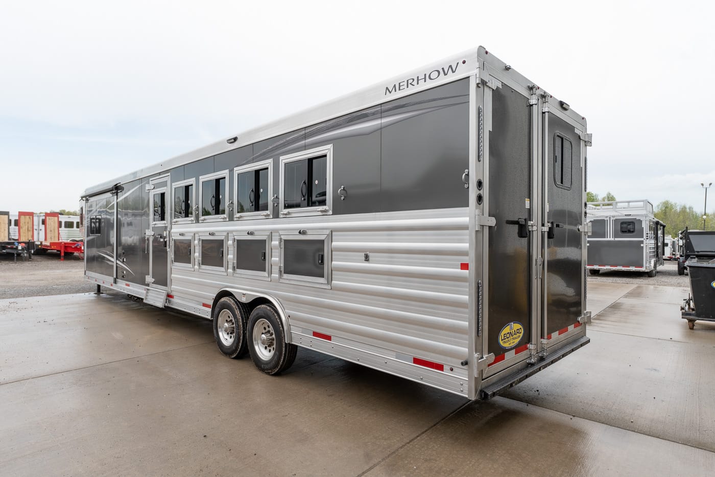 Silver Merhow horse trailer with multiple side windows and built-in storage compartments along the lower panel, parked on a lot.