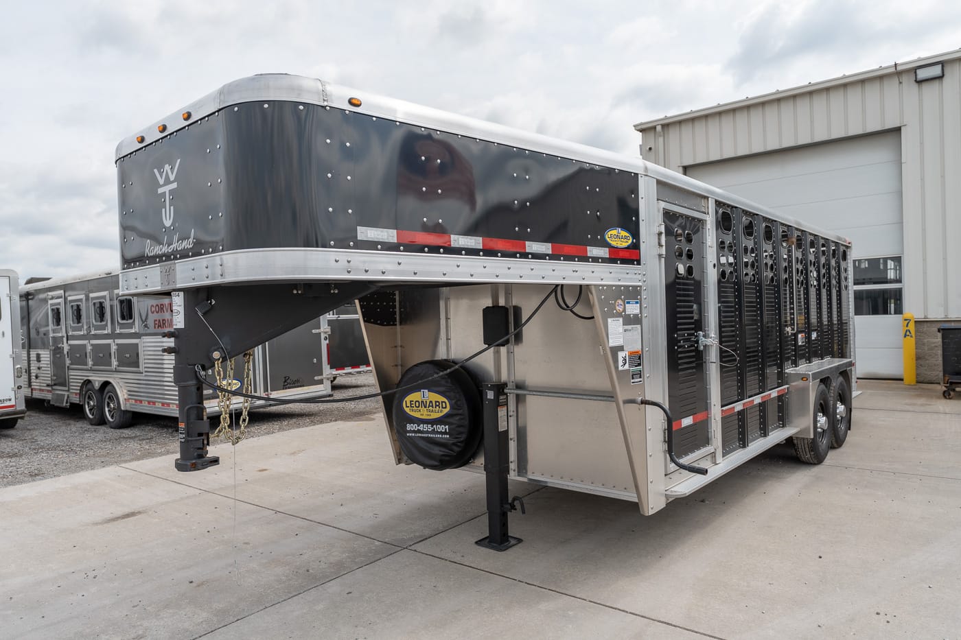 Silver and black gooseneck livestock trailer with ribbed vents, parked on a concrete lot beside other trailers; Leonard branding visible on the wheel cover and side panels.