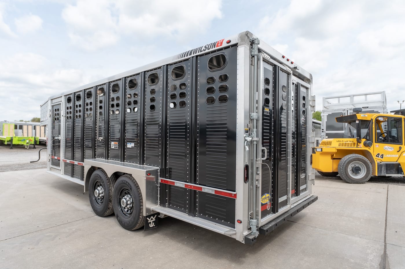 Livestock trailer with ventilation slots on a dual-axle aluminum body parked in a loading yard; a yellow forklift is in the background.