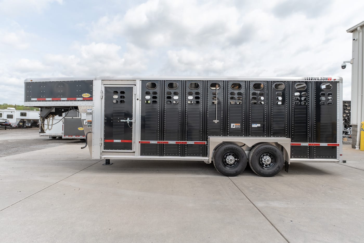 Large black multi-stall horse trailer with vented doors, parked on a concrete lot beneath a cloudy sky.