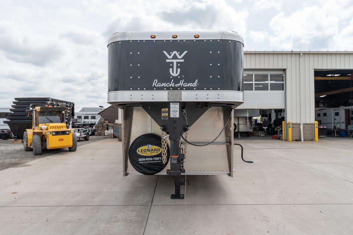 Front view of a black Ranch Hand gooseneck horse trailer parked in a repair yard, with a spare tire cover and forklift nearby