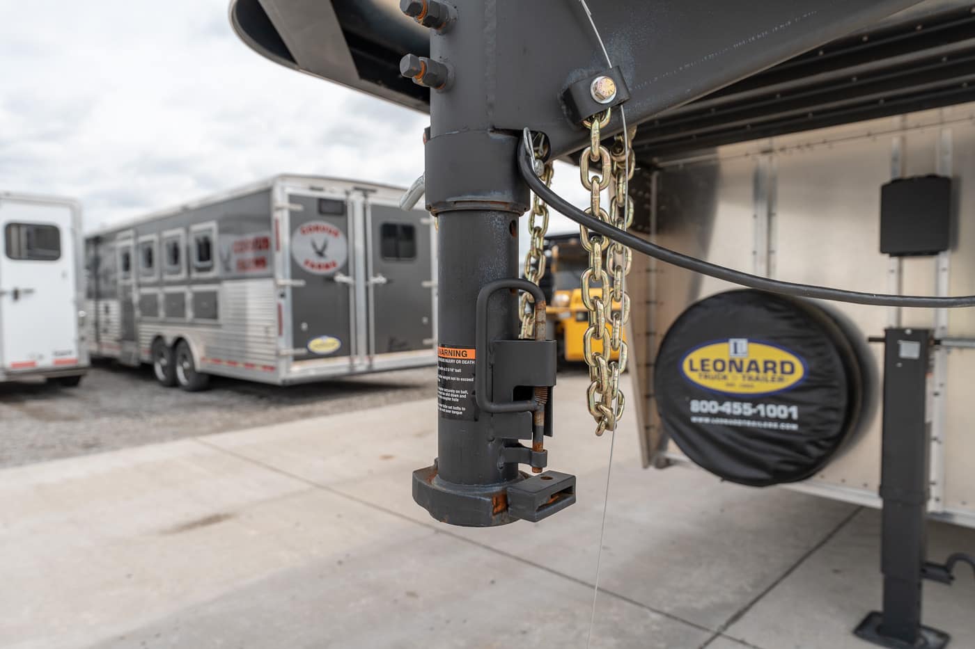 Close-up of a trailer hitch with a padlock and safety chains on a parked gooseneck trailer, with a Leonard spare tire and other trailers in the background.