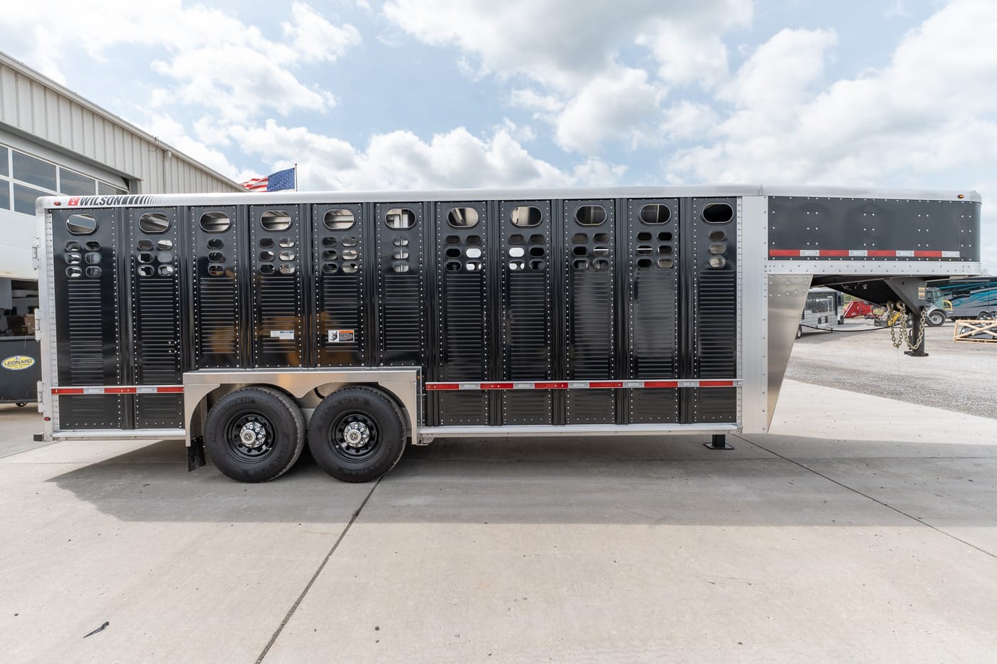 Black livestock trailer with vertical ventilation slats and twin rear wheels, parked on a concrete lot under a cloudy sky
