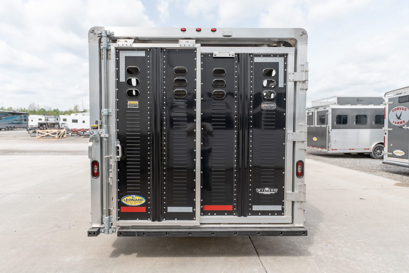 Rear view of a black-rated trailer door panel with vent slits and riveted metal framing, parked in a lot with other trailers nearby.