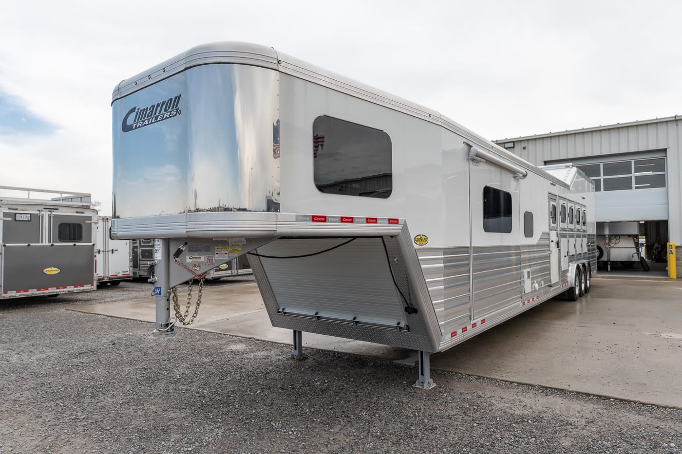 Large silver gooseneck horse trailer parked in a lot with other trailers and a warehouse in the background.