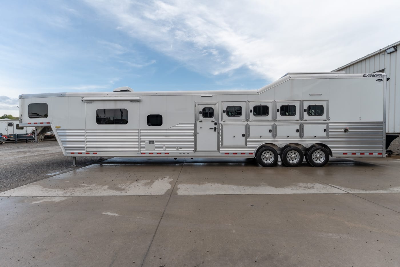 Long white aluminum horse trailer with multiple ventilation stalls and three axles parked on a concrete lot under a blue sky.