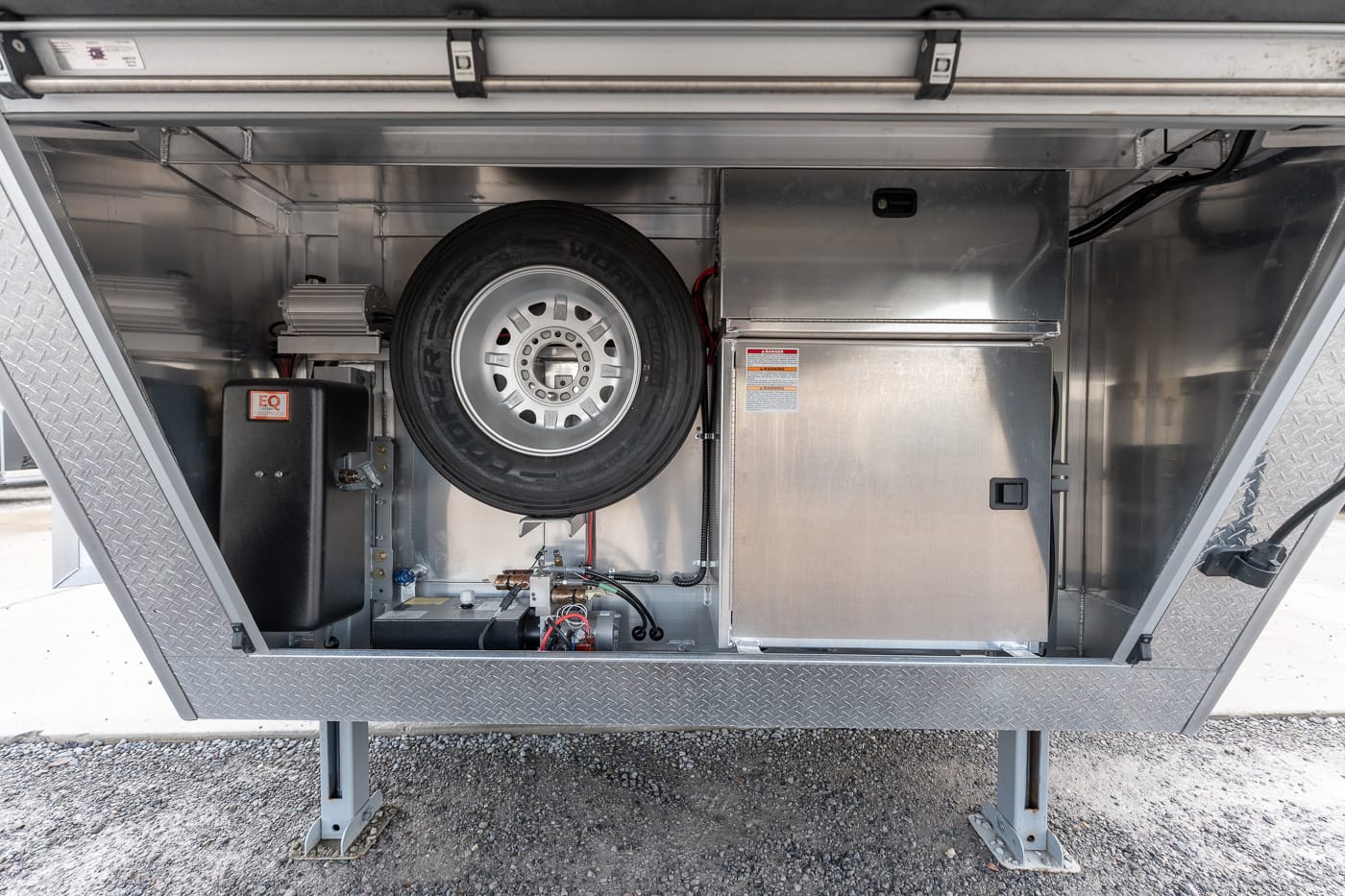Open equipment compartment of a utility trailer showing a mounted spare tire, battery box, and metal storage cabinet inside a diamond-plate frame nearby the landing legs