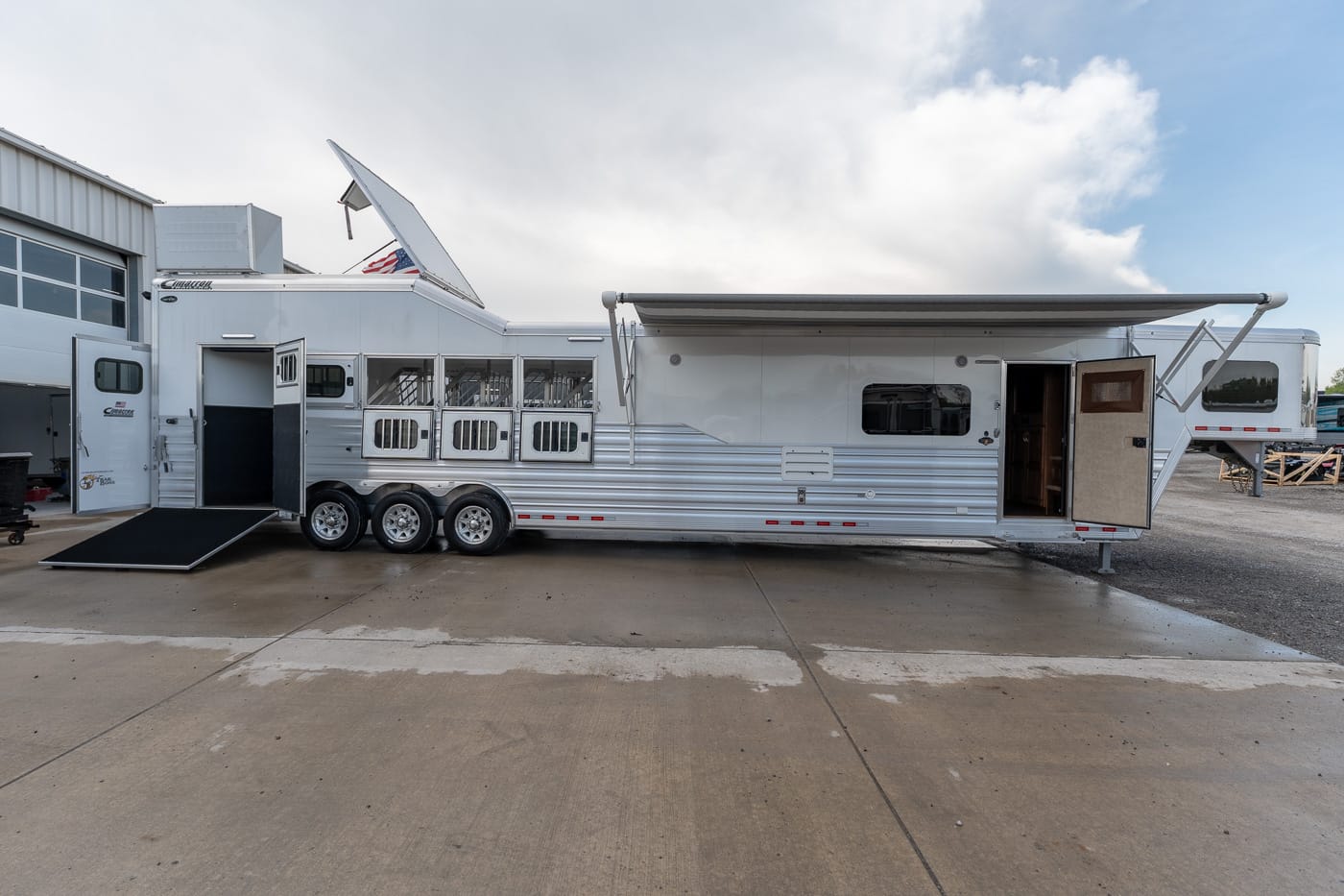 White long horse trailer with a lowered ramp and open side door on a concrete lot, plus an extended awning on the right side.