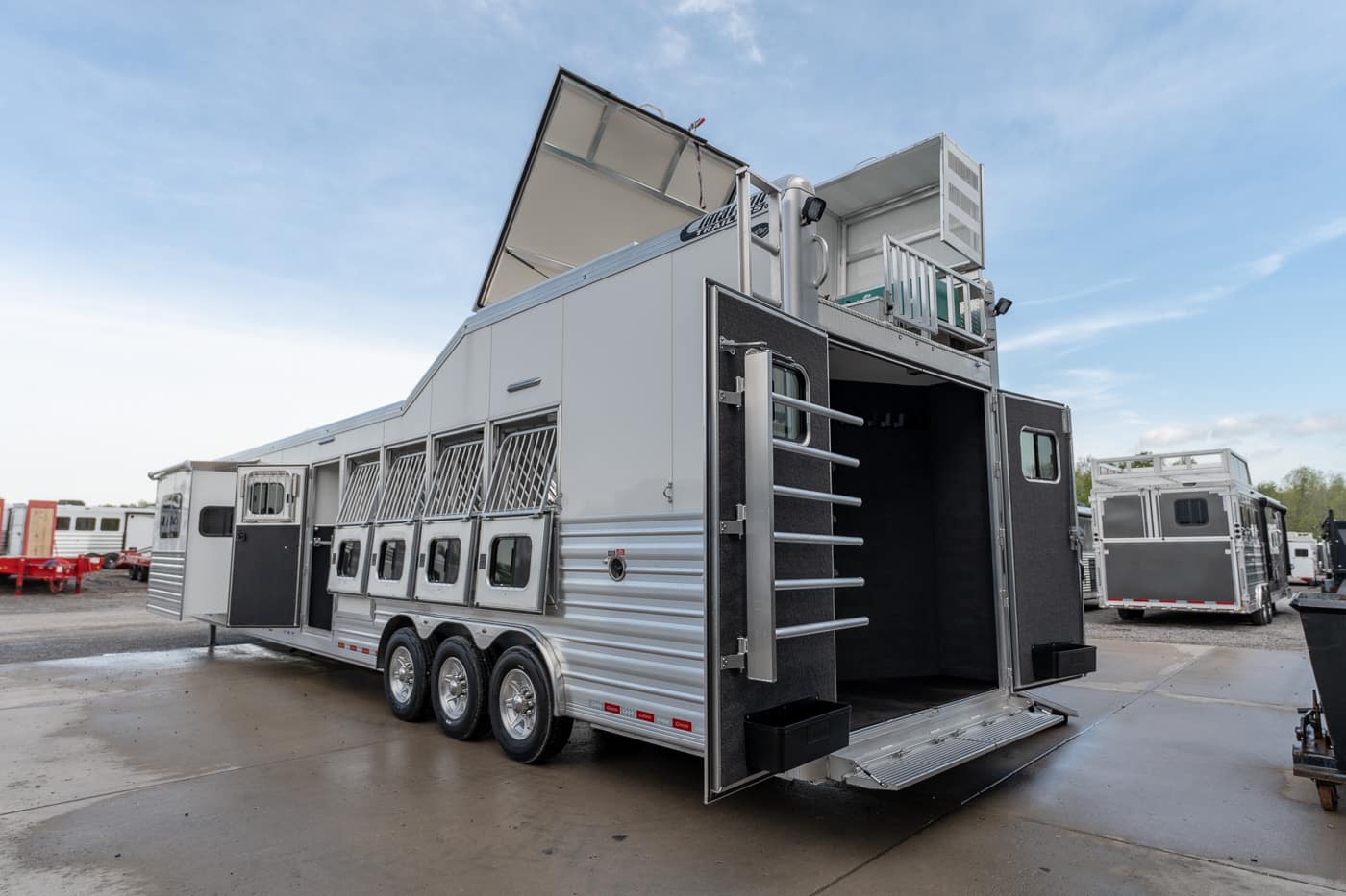 Silver multi-compartment horse trailer with three axles and the rear ramp down, exposing interior stalls and tack areas.