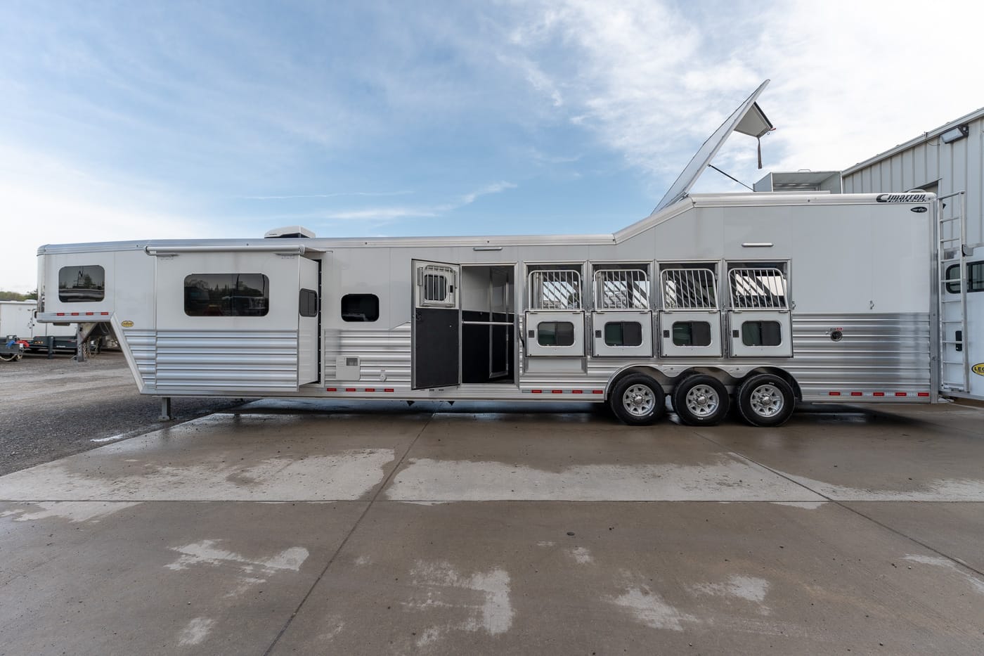 Long white horse trailer with multiple stall doors and ventilation bars parked on a concrete lot, under a blue sky.