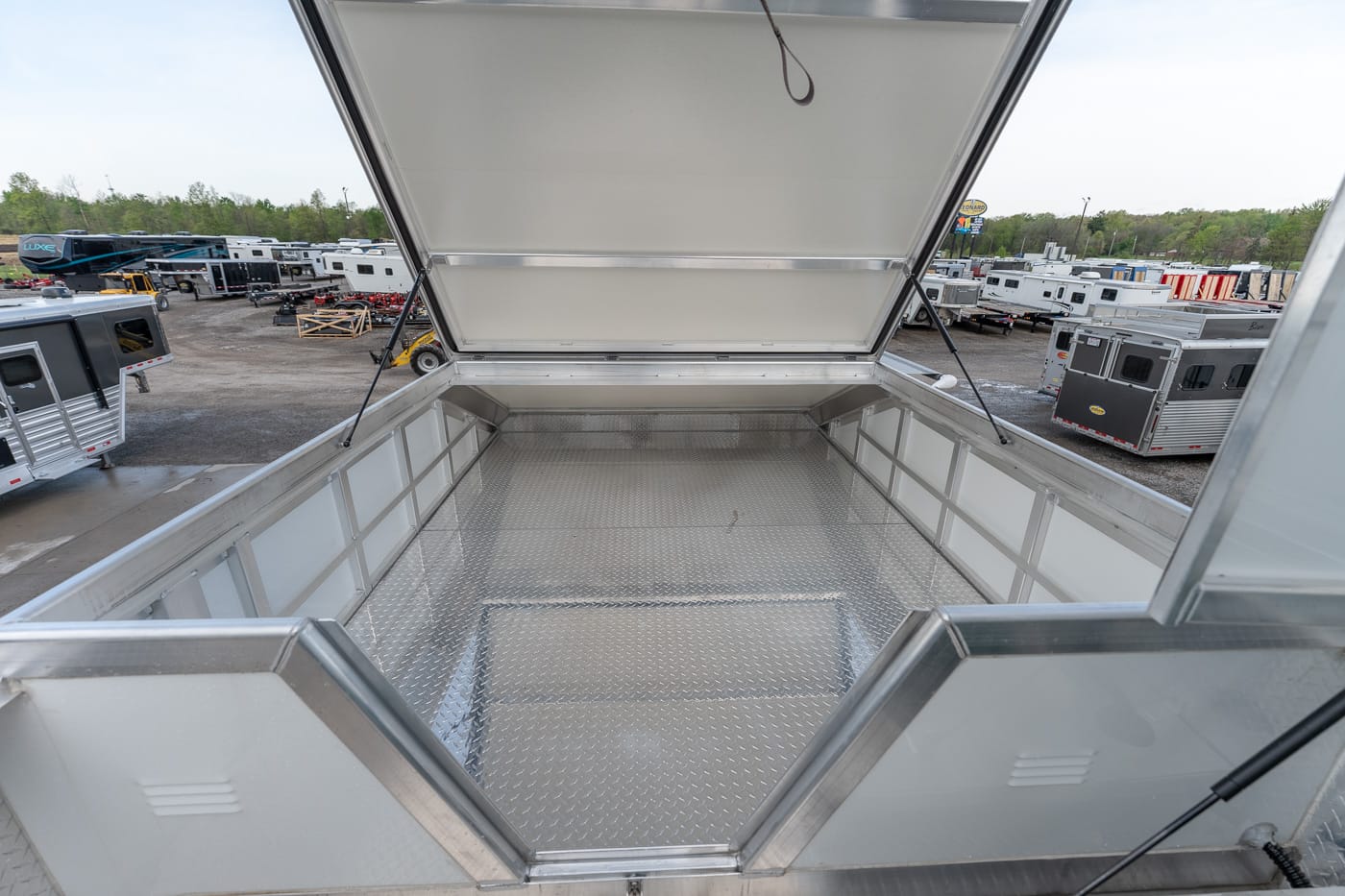 Open aluminum cargo trailer bed with a diamond-plate floor, parked among RVs and trailers in a lot.