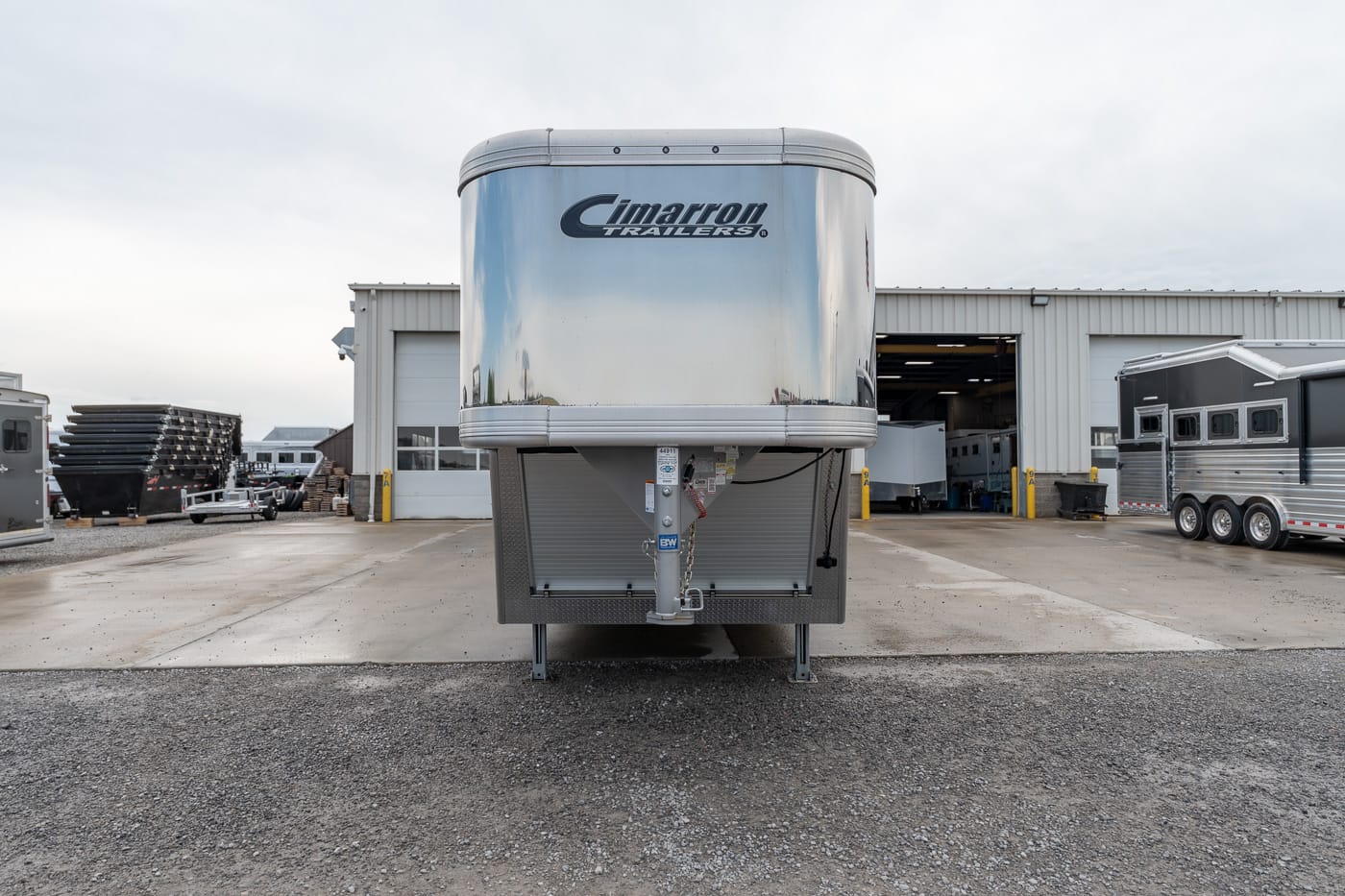 Front view of a shiny Cimarron gooseneck trailer parked on a gravel lot outside a service shop, with other trailers nearby.