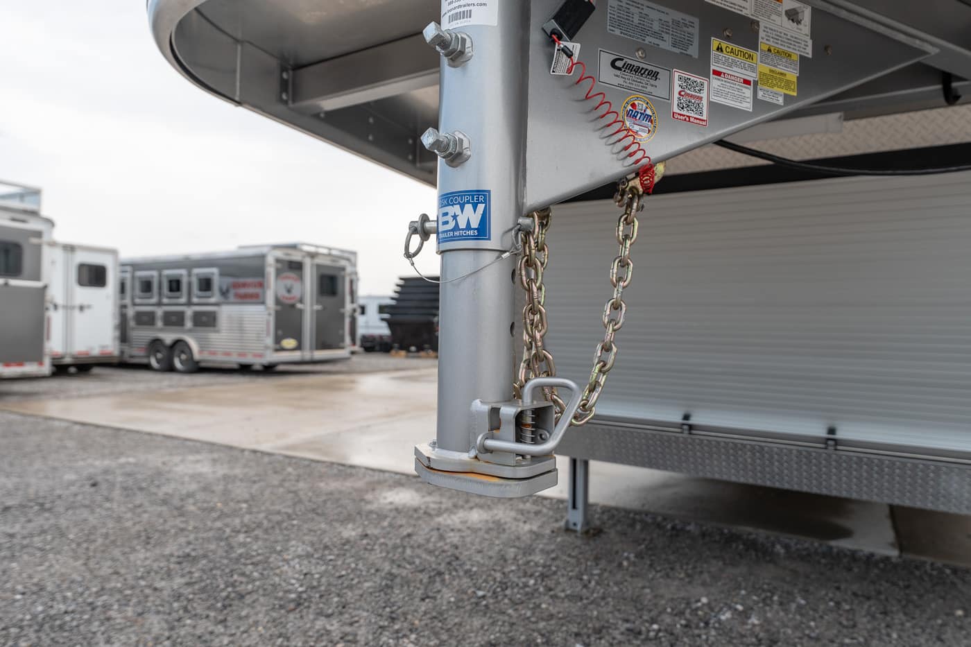 Close-up of a metal trailer hitch with safety chains and a latch, on a gravel lot with other trailers in the background.